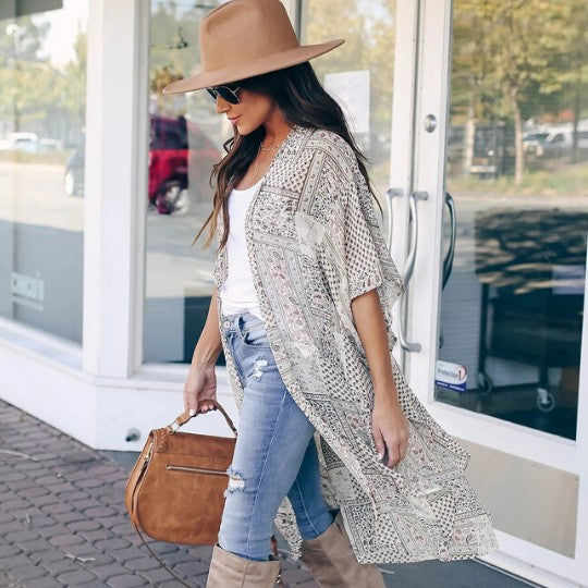 Woman wearing a patterned kimono, jeans, and boots with a brown hat and bag, standing outside a store.