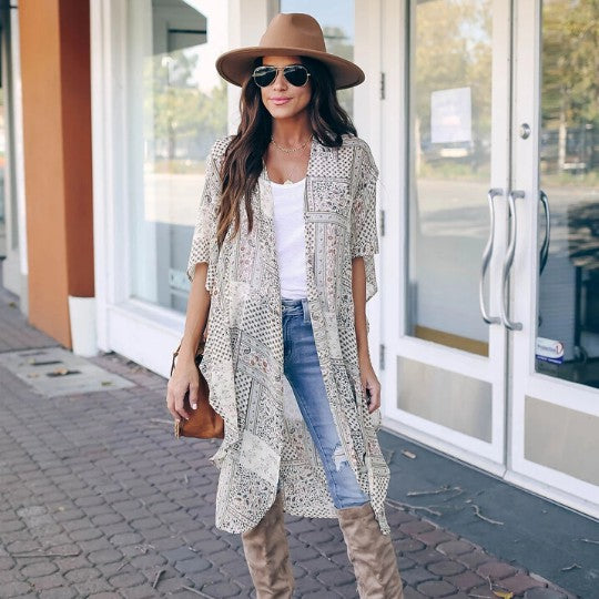 Woman wearing a patterned kimono, jeans, and boots with a brown hat and bag, standing outside a store.